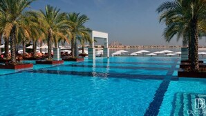 A swimming pool with palm trees and umbrellas, with a view of the city in the background. - Jumeirah Zabeel Saray Royal Residence Lagoon Villa (Dubai)
