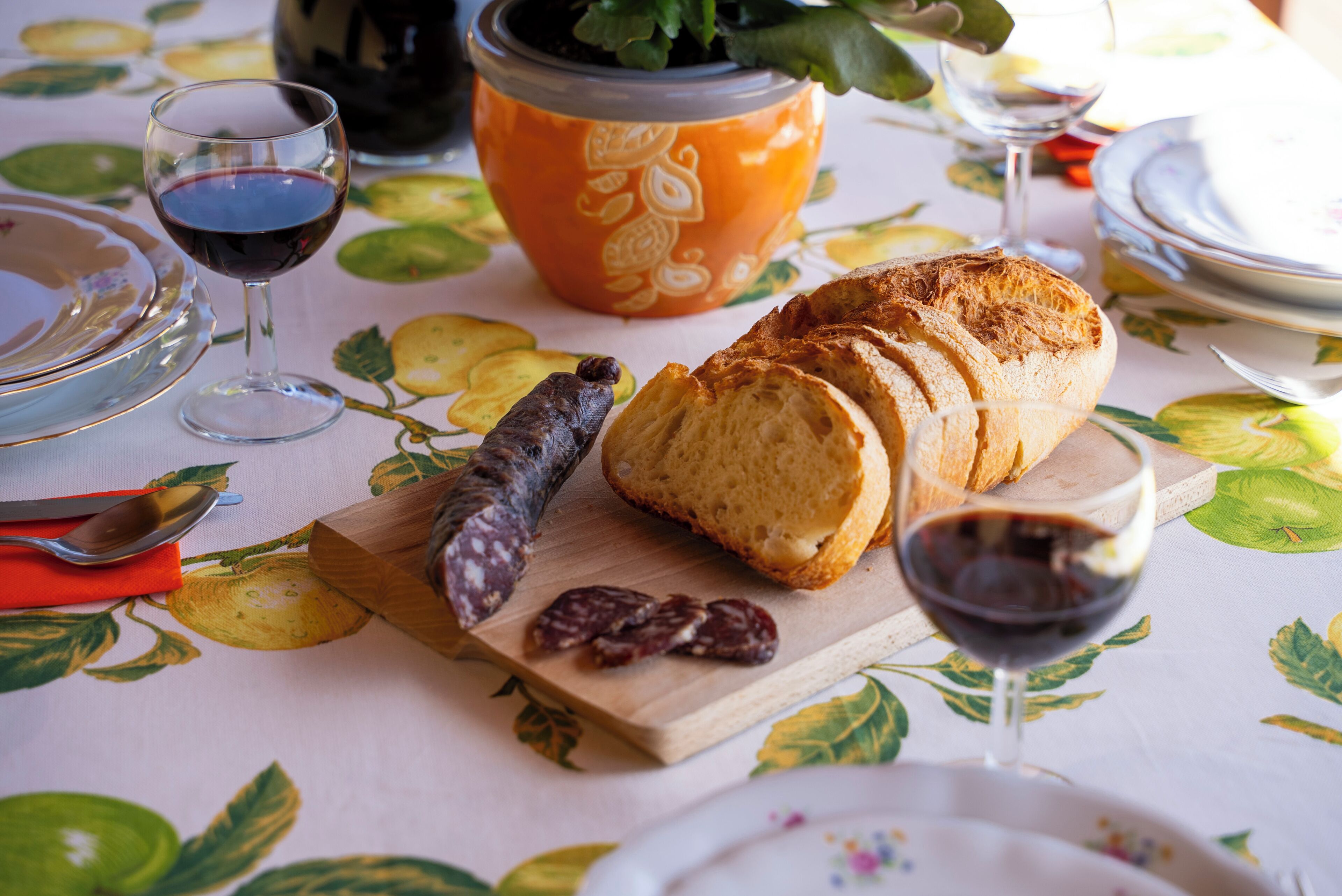 Ein Tisch mit einer blumigen Tischdecke, einem Brotlaib, einer Weinflasche und einem Holzer Schneidebrett mit Brotscheiben und einer Scheibe Salami.