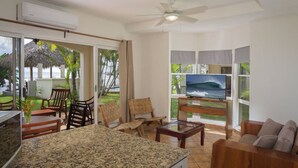 A modern living room with a TV, wooden furniture, a ceiling fan, and large windows overlooking a garden.