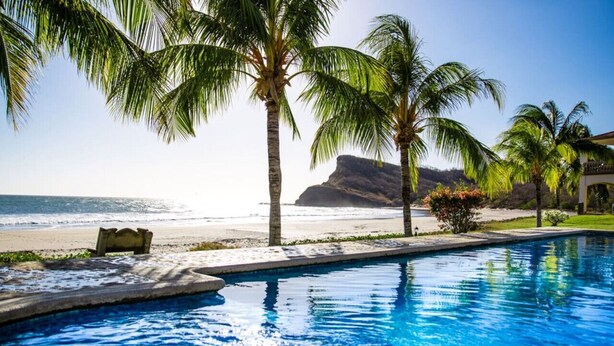 A swimming pool with clear blue water, surrounded by palm trees and a view of the beach and ocean.