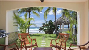 A patio with wooden chairs and a table, overlooking a beach with palm trees and a thatched-roof structure.