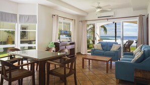 A spacious living area with a large window overlooking the ocean, a blue sofa, a dining table with chairs, and a ceiling fan.