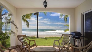 A covered patio with wooden chairs and a table, overlooking a beach with palm trees and the ocean.