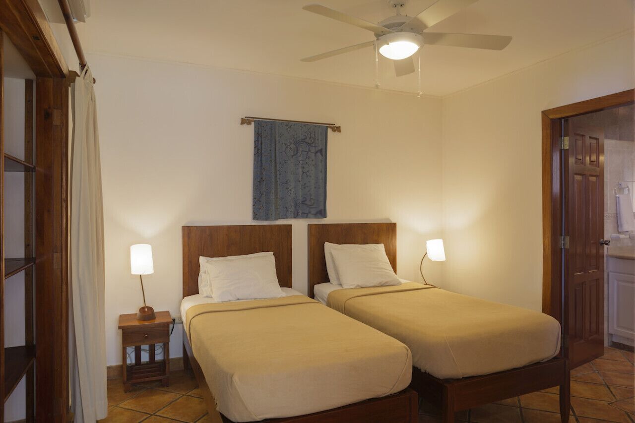 Une chambre d’hôtel avec deux lits simples, un ventilateur de plafond, une petite table avec une lampe et une porte en bois.