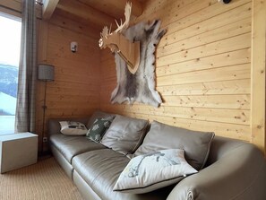 Interior de una cabaña de madera con un sofá de cuero, cojines decorativos y un trofeo de piel de animal en la pared.