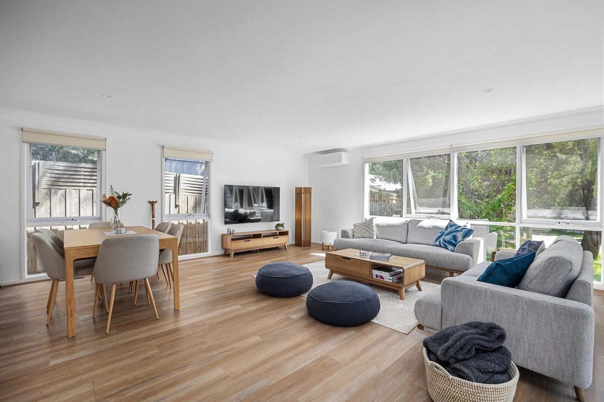 A modern living room with a dining area, large windows, and a view of greenery.