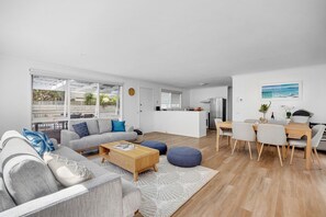 A modern living room with a sofa, coffee table, and ottoman, a dining area with chairs, and a kitchen in the background. - Allure Beach House Rye. (Rye)