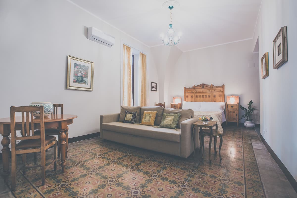 Quarto com sofá, uma cadeira de madeira, mesa pequena, mesa de cabeceira com abajur, cabeceira de madeira, piso estampado e luminária no teto.
