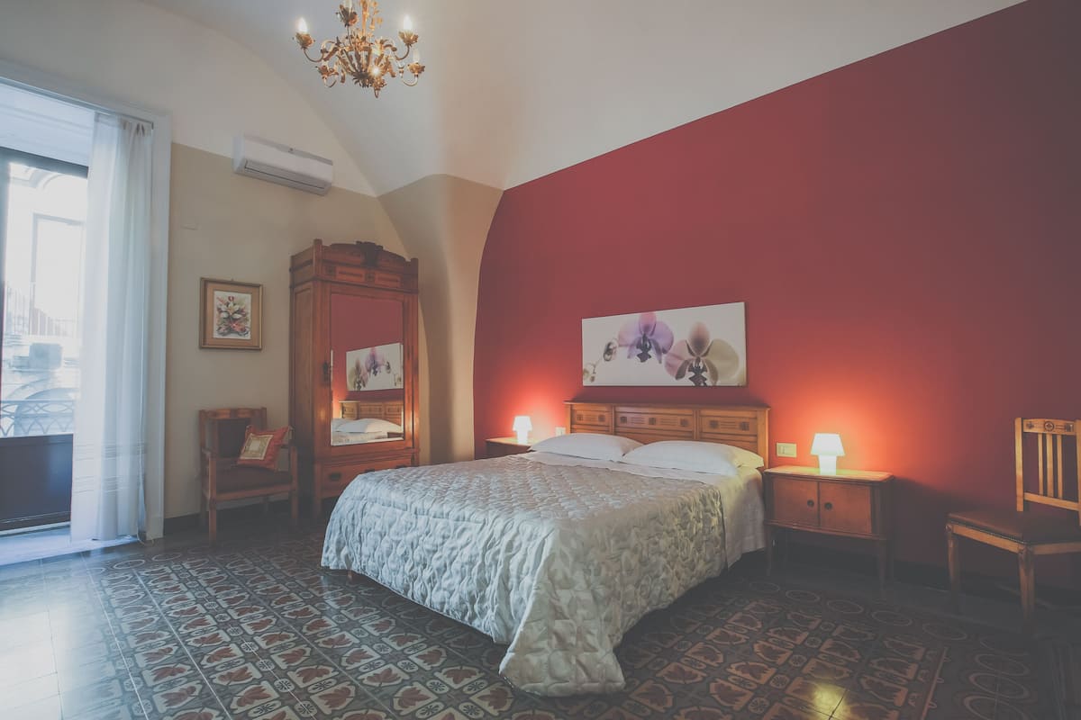 Quarto com cama, mesas de cabeceira, uma cadeira de madeira, um lustre e uma janela grande com cortinas.