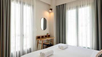A hotel room with a bed, a desk, a chair, a mirror, and sheer curtains.