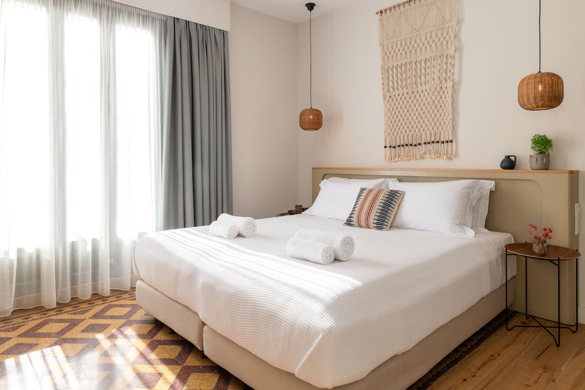 A neatly made bed with white linens and two rolled towels, a bedside table with a vase of flowers, and a macrame wall hanging.