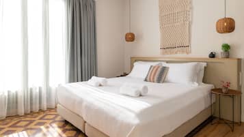 A neatly made bed with white linens and two rolled towels, a bedside table with a vase of flowers, and a macrame wall hanging.