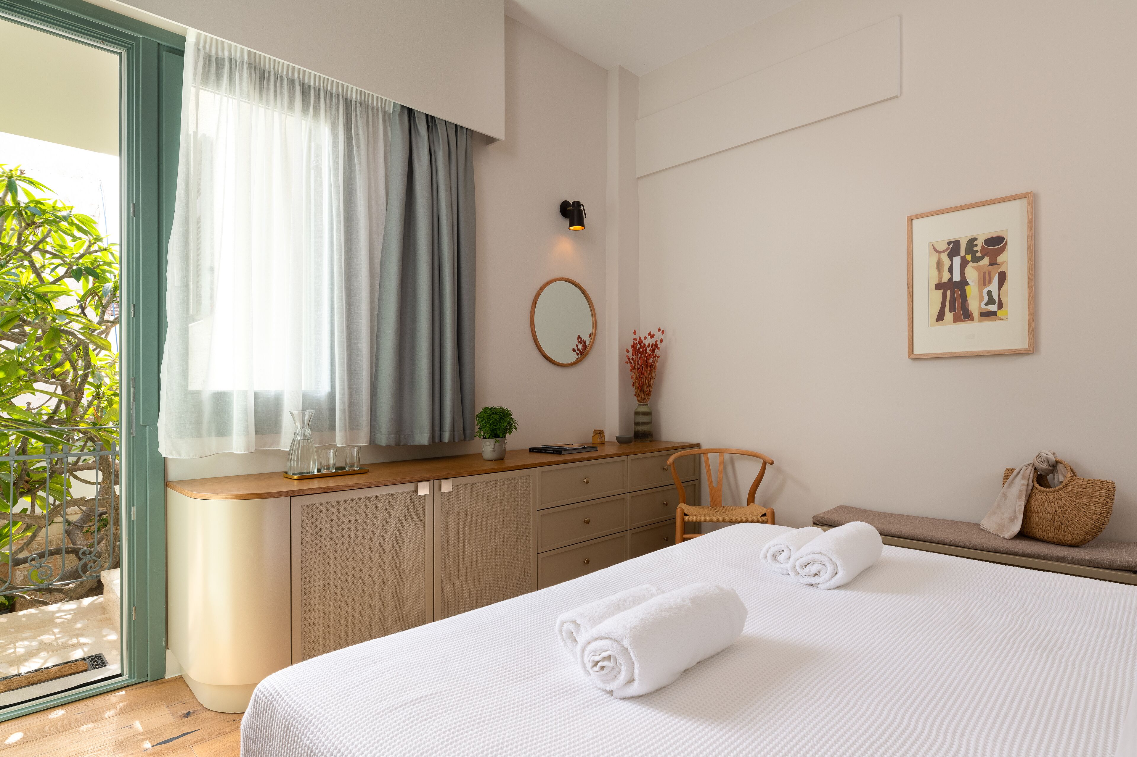 A hotel room with a bed, a desk, a chair, a window with curtains, a wall-mounted lamp, a framed picture, and a small plant.