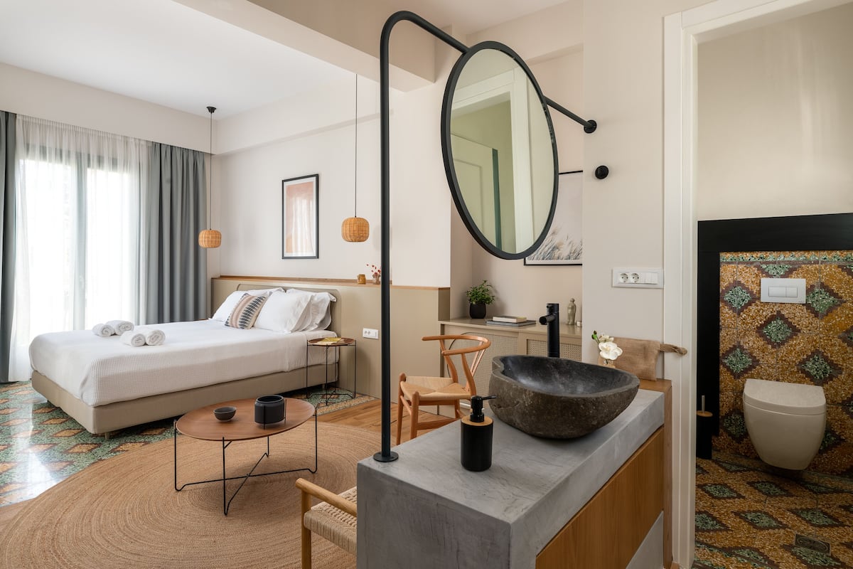 A modern hotel room with a bed, a round table, a mirror, a sink, and a bathroom.