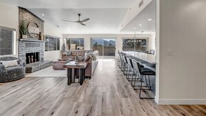 A spacious living area with a fireplace, a large sofa, a dining table with bar stools, and a view of mountains through large windows. - Prime Location, Walk To Restaurants, Enjoy The Sunrise And Red Rock Views (Sedona)