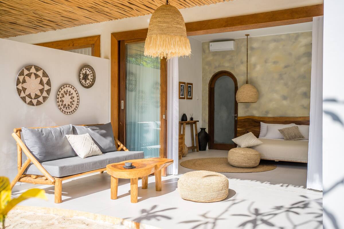 Una habitación con sofá de madera, una mesa de madera, un pouf tejido y una lámpara colgante.