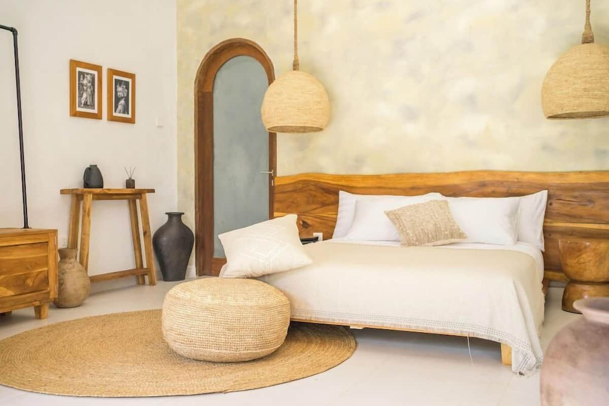 Un dormitorio con cabecera de madera, un reposapiés redondo tejido, una mesita de noche con un jarrón y dos lámparas colgantes.