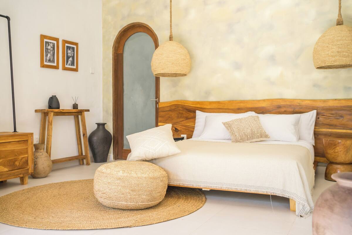 Un dormitorio con cabecera de madera, un taburete redondo tejido, un tapete redondo tejido, una mesita de noche con un jarrón y dos cuadros en la pared.