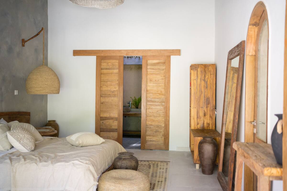 Un dormitorio con cama, mesitas de noche, lámpara colgante, espejo y puertas de madera que dan a un baño.