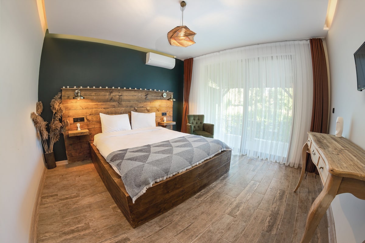 A bedroom with a wooden headboard, a large bed, a wooden desk, and a window with sheer curtains.