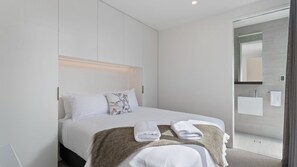 Een moderne hotelkamer met een bed, nachtkastje en een badkamer met een wastafel en spiegel.