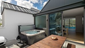 Een moderne buitenjacuzzi met een houten tafel en barbecue, grote schuifdeuren die leiden naar een eethoek met uitzicht op een heuvel.