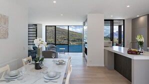 A modern kitchen with a dining area, large windows with a view of a mountainous landscape, and a small island with fruit and flowers.