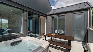 A modern outdoor hot tub area with a wooden table and benches, large sliding glass doors, and a view of a natural landscape.