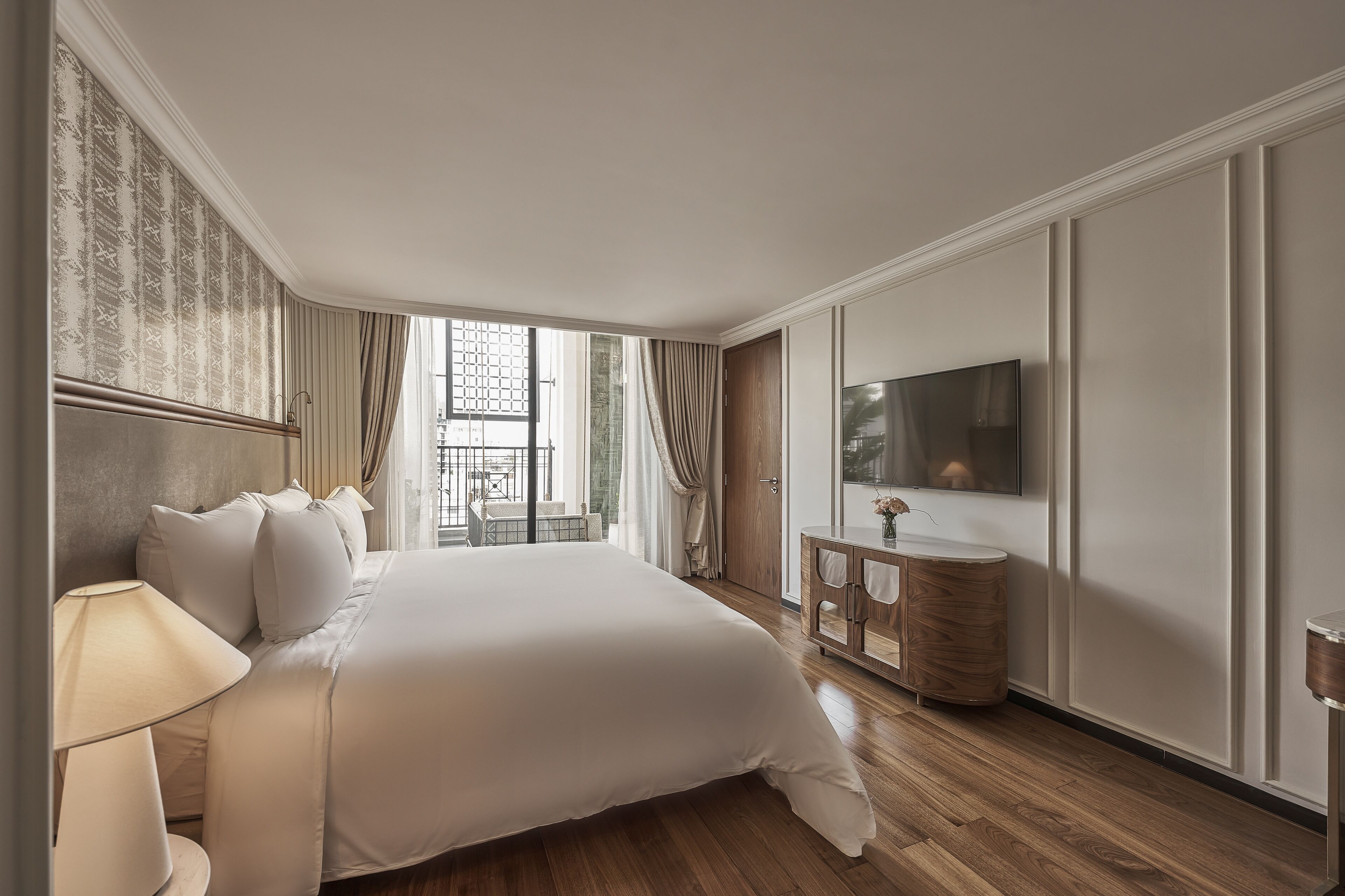 A hotel room with a large bed, a TV, a wooden floor, and a balcony with curtains.