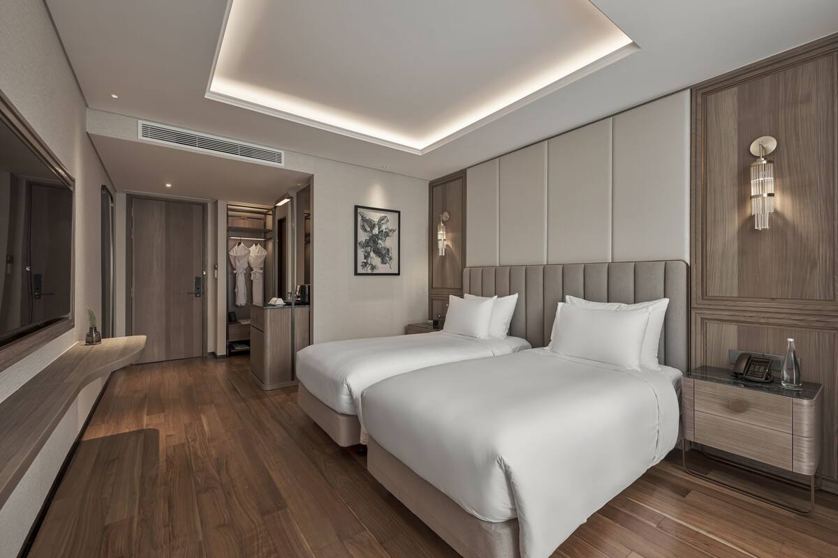 Ein modernes Hotelzimmer mit zwei Betten, einem Flachbildfernseher, einem Nachttisch mit Lampe und einem Kleiderschrank.
