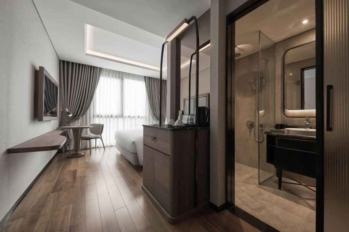 Habitación de hotel moderna con piso de madera, un escritorio con silla, un espejo grande, zona de ducha y una ventana con cortinas.