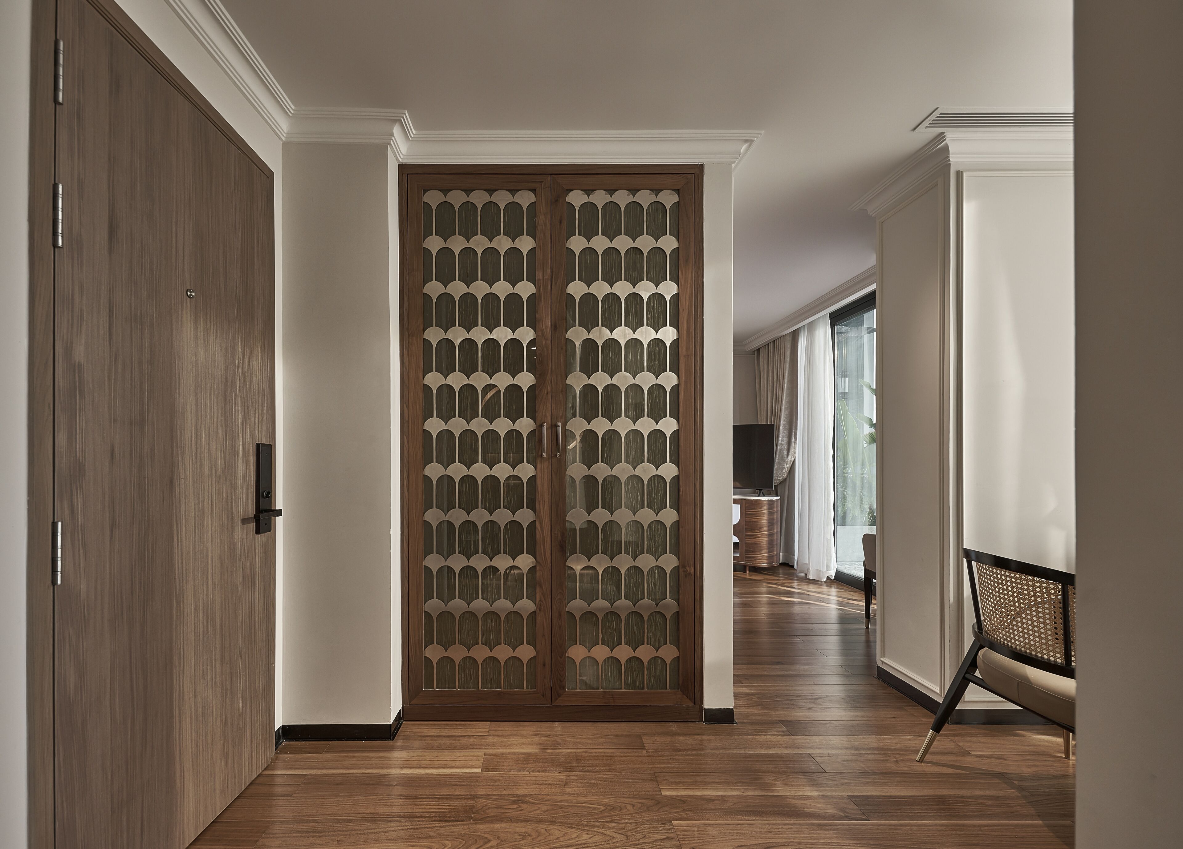 A hallway with a wooden floor, a sliding door with a geometric pattern, and a view into a room with a television and a chair.