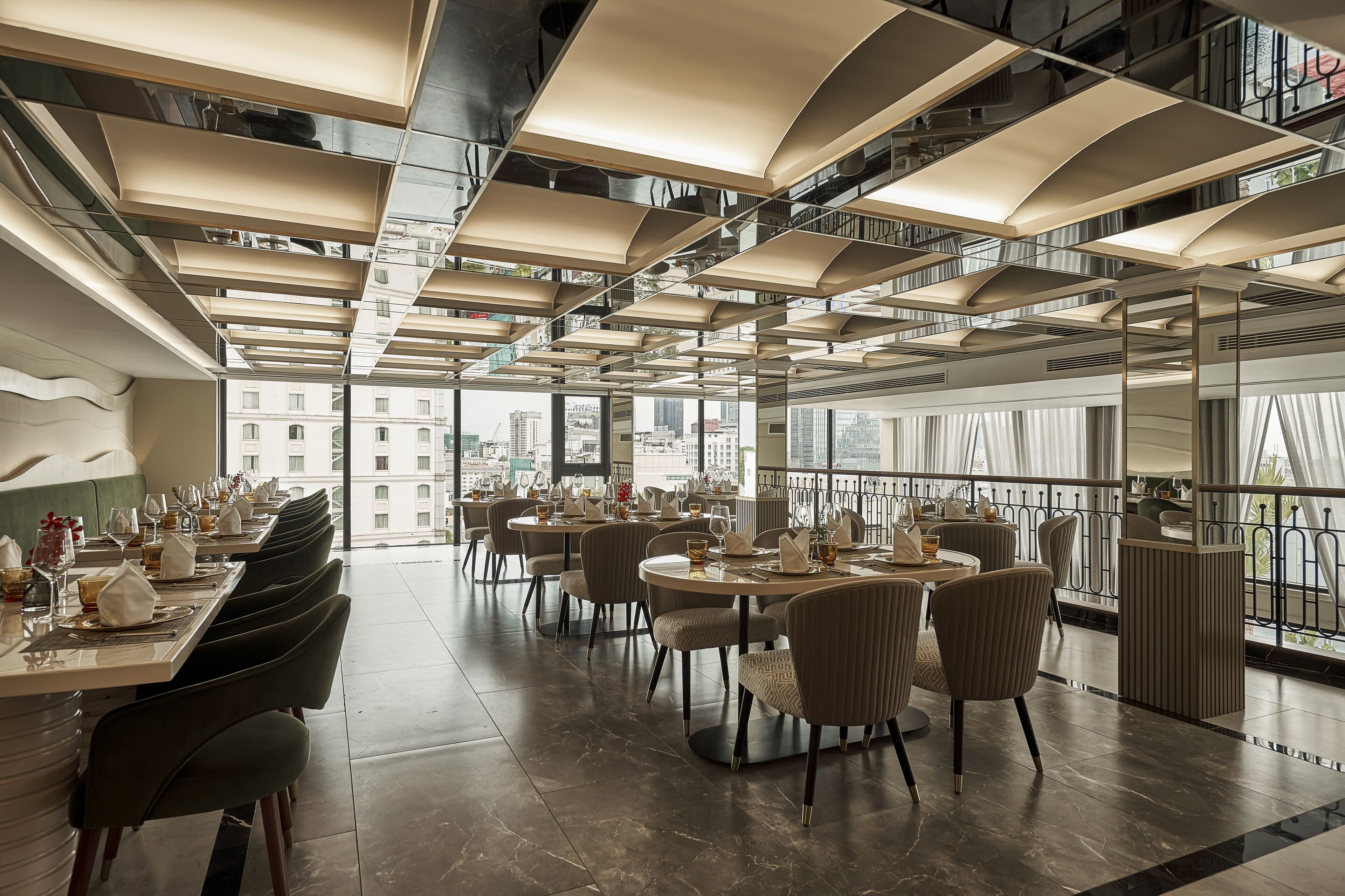 A modern dining area with multiple tables and chairs, large windows offering city views, and a high ceiling with exposed ductwork.