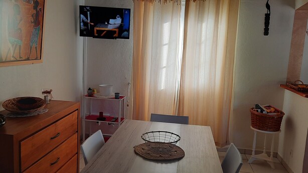 En matplats med ett träbord, stolar, en tv som är monterad på väggen, ett fönster med gardiner och en liten hylla med en korg.