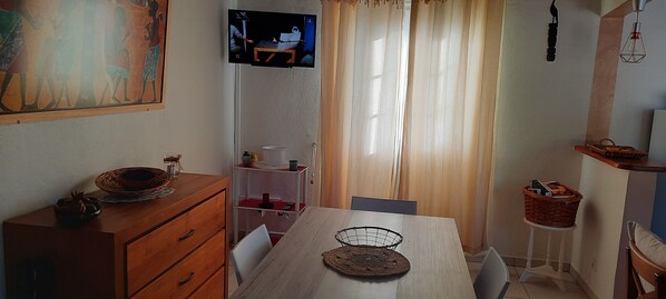 Un comedor con una mesa de madera, sillas, un televisor montado en la pared, una ventana con cortinas y un estante pequeño con una cesta.