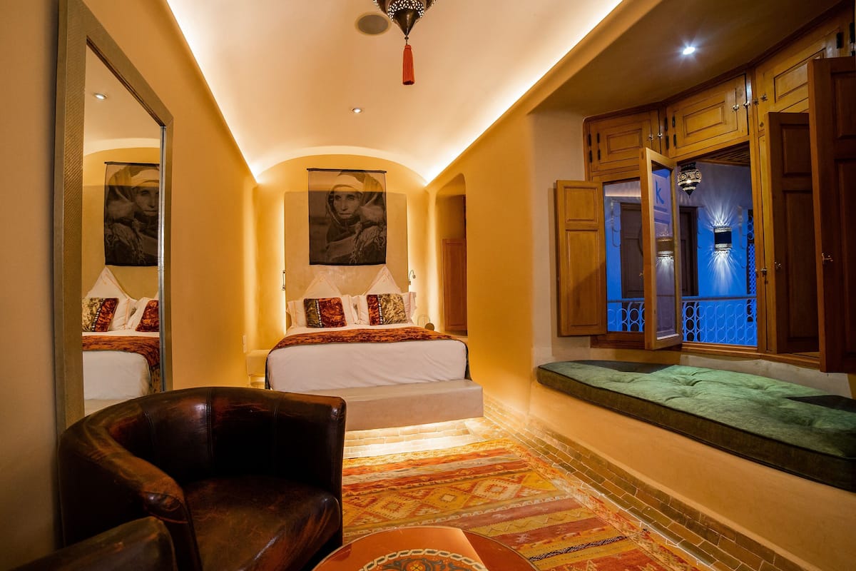Habitación de hotel con cama, una silla, un banco, un espejo y ventana con vista.