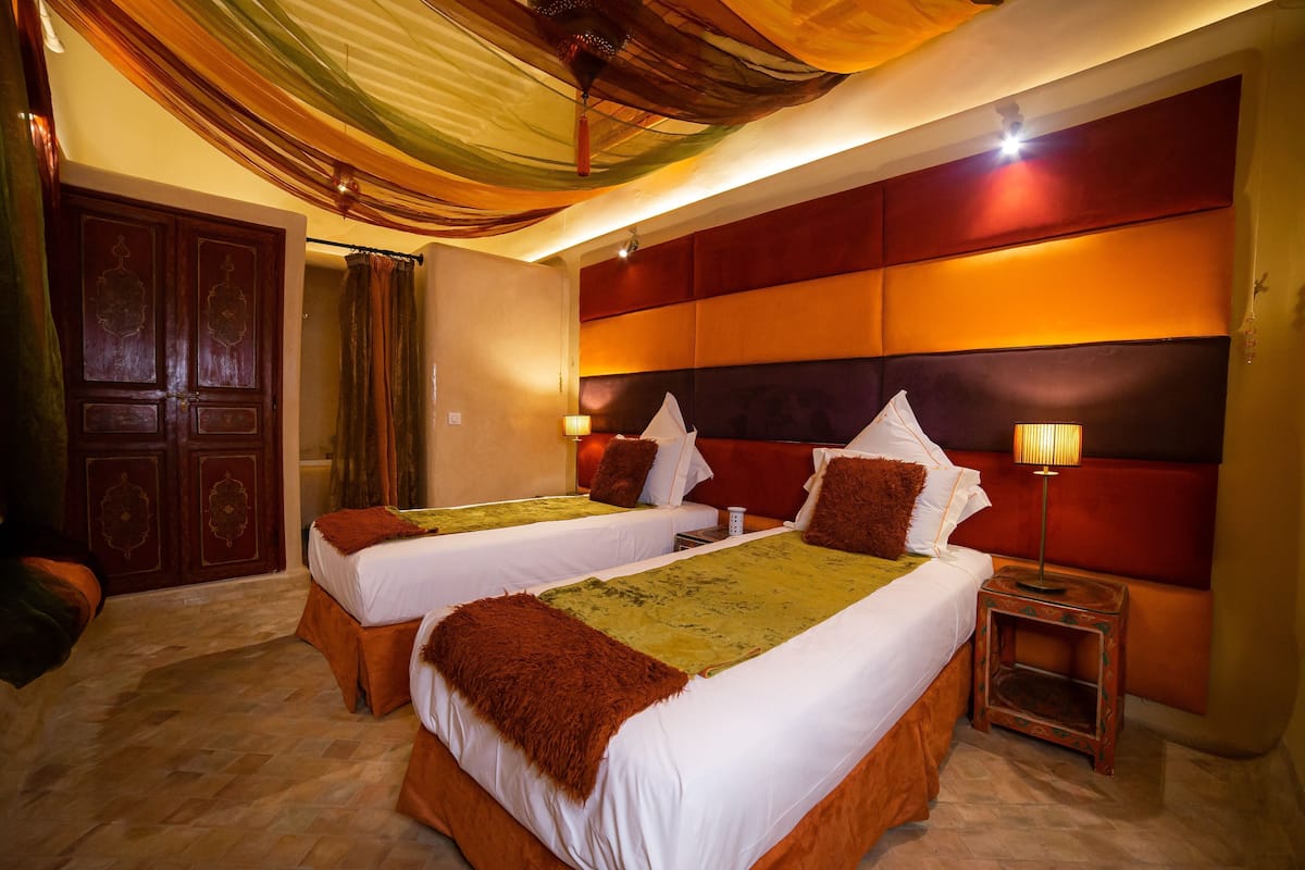 Habitación de hotel con dos camas, un armario de madera, una mesita de noche con lámpara y techo decorativo.