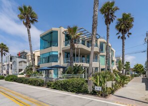 A modern multi-story building with balconies, surrounded by palm trees and greenery.