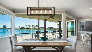 Un espace repas offrant une vue sur la piscine et la plage, avec une table en bois, des chaises et un luminaire suspendu.