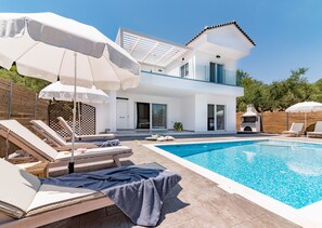 Una casa moderna de dos pisos con piscina, sillas de descanso y sombrillas.