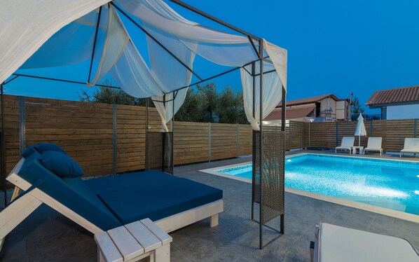 Un área junto a la piscina con una zona de estar cubierta, piscina y valla de madera.