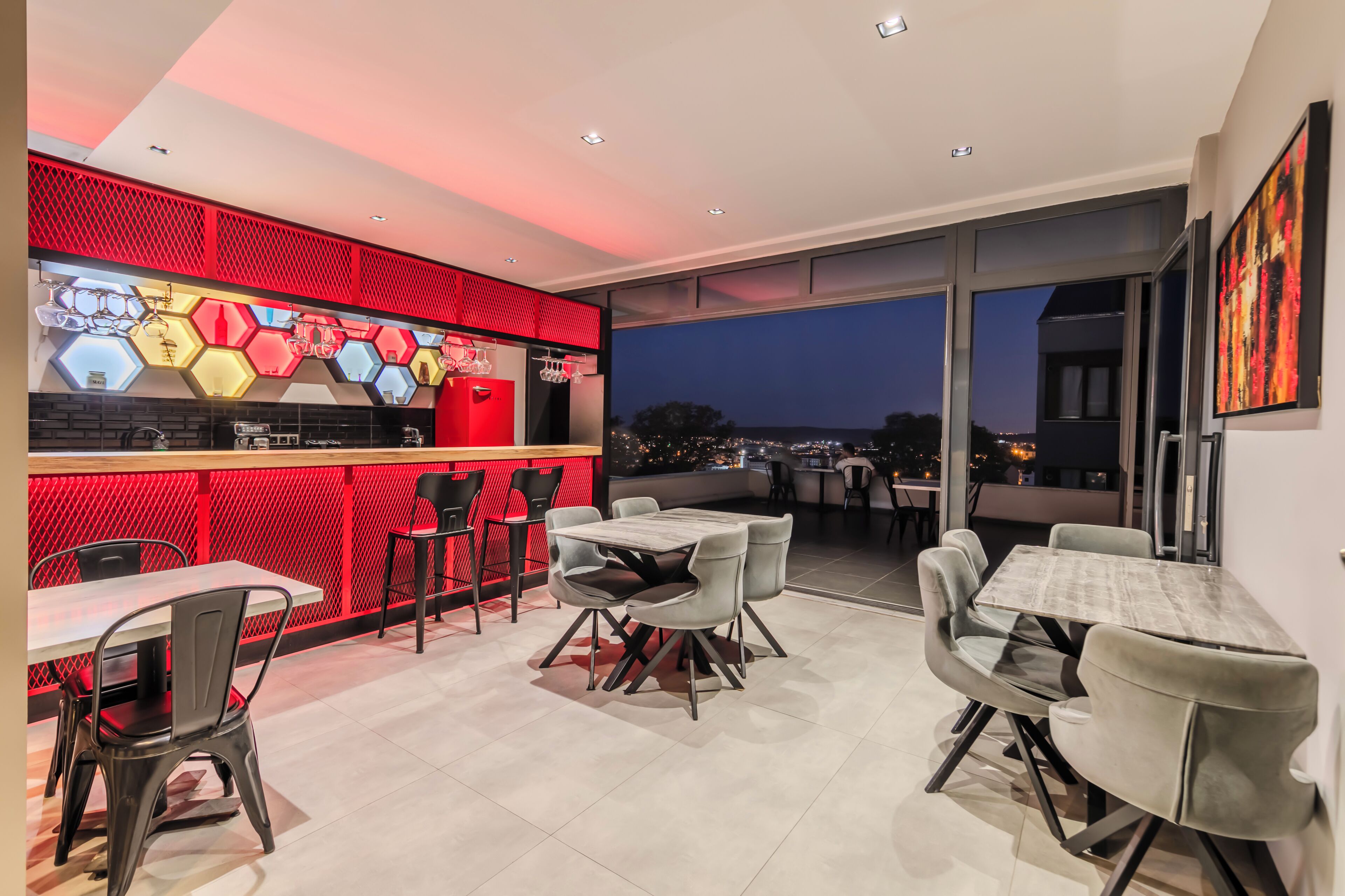 Un comedor moderno con una pared de acento roja, lámparas hexagonales y vistas de la ciudad al atardecer.