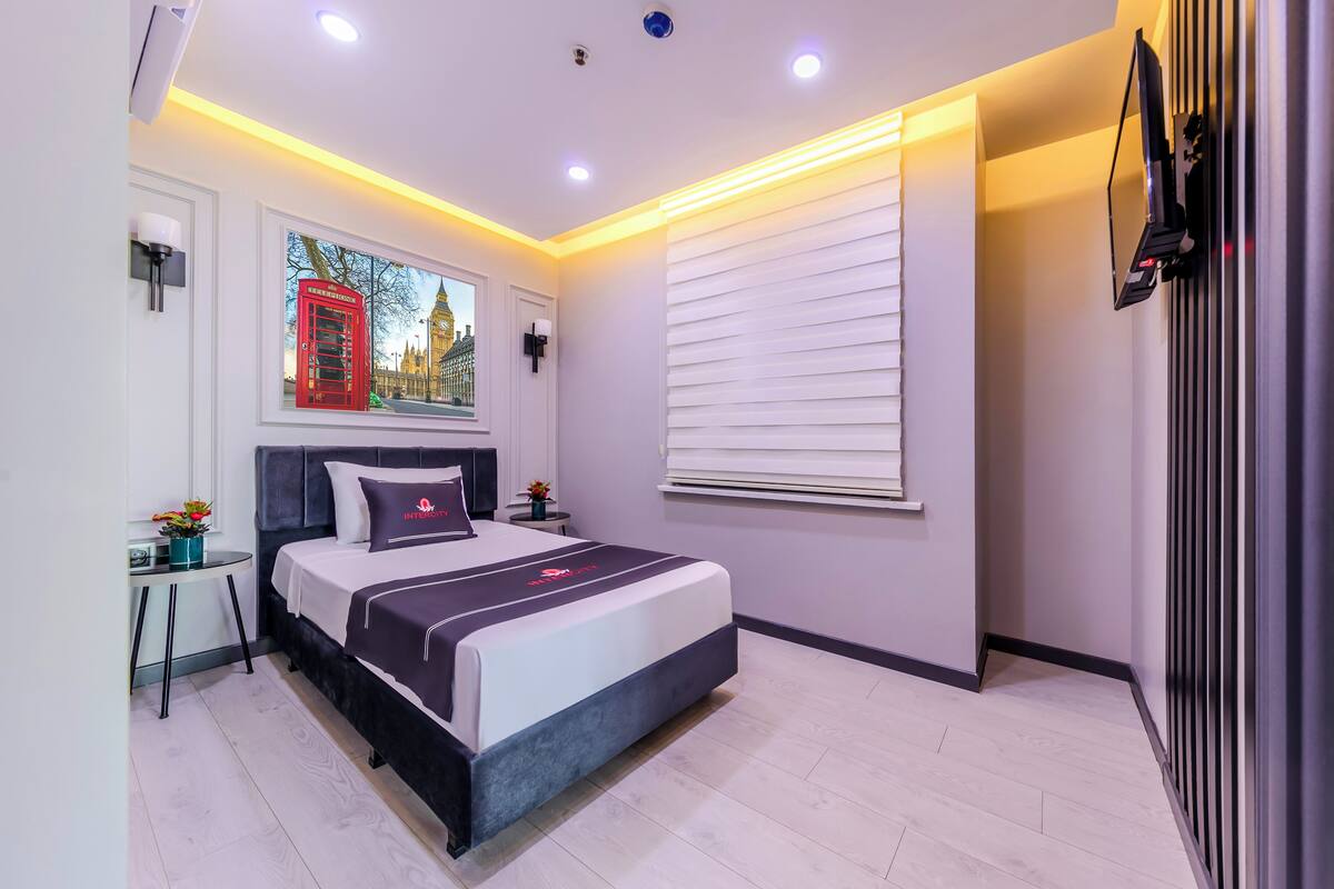 A modern bedroom with a bed, bedside tables, a window with blinds, a wall-mounted TV, and a painting of a cityscape.