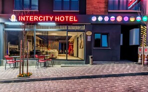 L’extérieur de l’hôtel Intercity présente une façade moderne, une entrée en verre et un espace extérieur aménagé avec des chaises et des tables rouges.