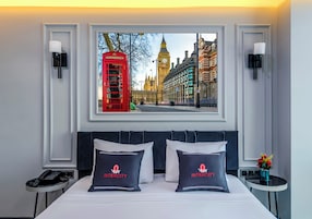 Habitación de hotel con una amplia ventana que ofrece vistas al Big Ben y un teléfono rojo, una cama bien hecha con dos almohadas etiquetadas como "INTERCITY", una mesita de noche con una lámpara y una pequeña mesa con un jarrón de flores.