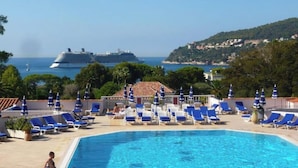 Une piscine bordée de chaises longues bleues et d’ombrelles, un grand navire de croisière en arrière-plan et une vue sur la côte.