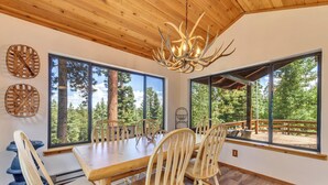 A dining area with a wooden table and chairs, large windows with a forest view, and a unique antler chandelier. - Charming Duck Creek Village Cabin | Fireplace (Duck Creek Village)
