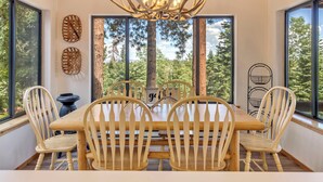 A dining area with a wooden table and chairs, a chandelier, and large windows with a forest view. - Charming Duck Creek Village Cabin | Fireplace (Duck Creek Village)