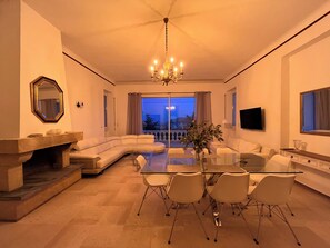 A modern living room with a glass dining table, white sofas, a stone fireplace, and a large window overlooking a balcony.