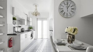 A modern kitchen with white cabinetry, a dining area with a white table, and a large wall clock. - The Roost Group - 1 Bedroom - Ground (Gravesend)